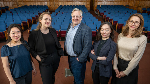 Five people pose for a group photo standing in front of a rows of auditorium seating